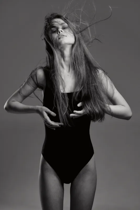 Black and white portrait of a young woman with flowing hair wearing a black bodysuit expressing emotion and movement in a studio setting.