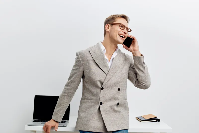 smiling young caucasian man talking smartphone double breasted blazer casual outfit standing near desk laptop digital nomad remote work concept