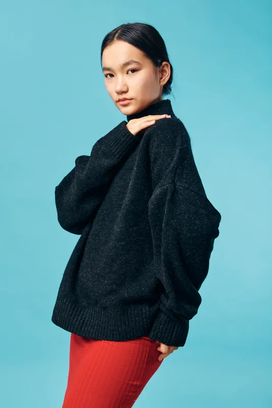 Young woman with long black hair wearing a black oversized sweater and red skirt posing against blue background with a soft confident expression and casual style.