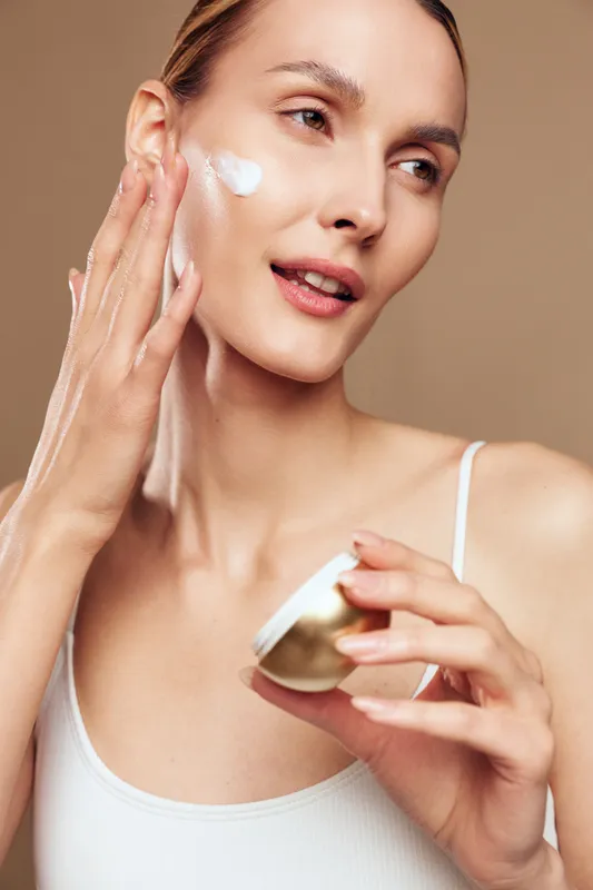 A young woman in a white tank top applies moisturizing cream to her face with her fingers. The neutral background highlights skincare and beauty care routine concepts.