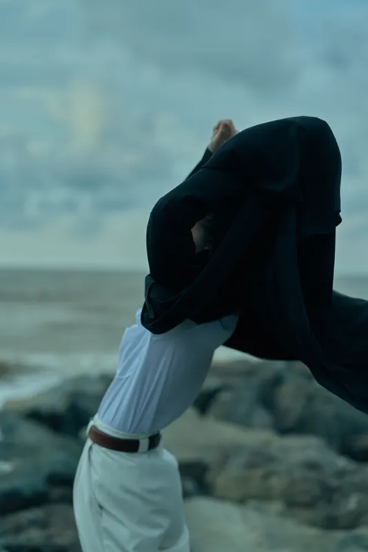 Movement, wind, fabric, garment, shore, ocean in background as white shirt and belt lift a dark cloak over rocky coast