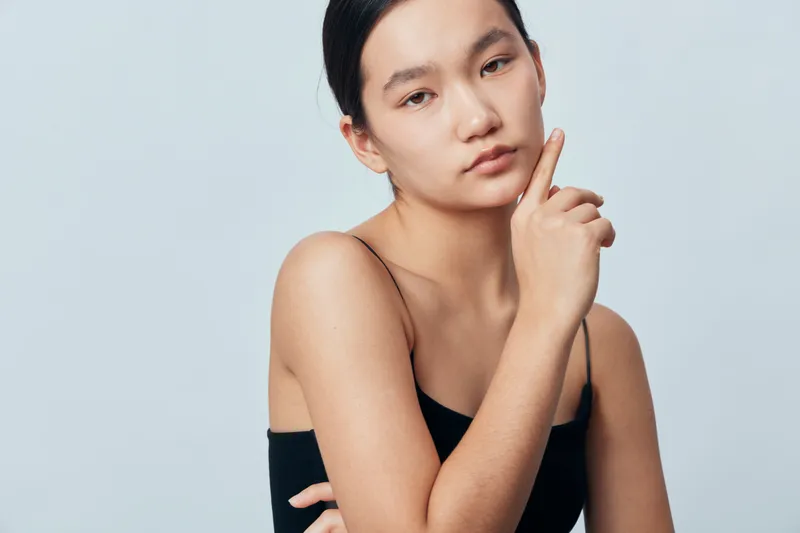 Young woman with natural skin and minimal makeup poses with hand near face looking thoughtful and confident against plain background in studio portrait. Beauty, fashion, and lifestyle concept.