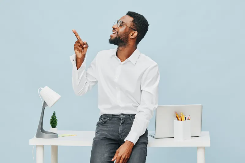 Smiling man with dark skin, wearing white shirt and dark jeans, sitting at modern office desk points upward with finger, looking away, cheerful expression, workspace with laptop, pen holder, and lamp 