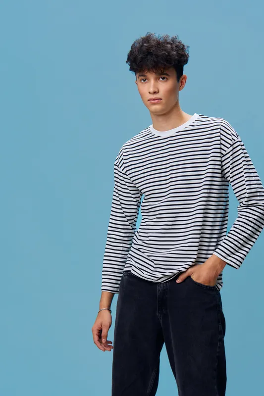 Young man wearing a striped long sleeve shirt and black jeans standing against a blue background with casual pose and curly hair in studio.