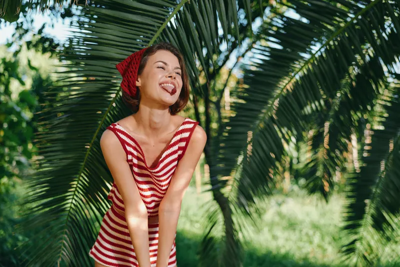 A cheerful young woman in a red and white striped dress smiles playfully among tropical palms, enjoying a sunny day outdoors, radiating joy and energy. Her bright expression and relaxed posture
