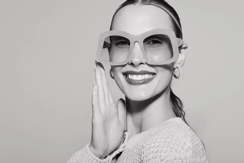 Happy white woman with fair skin and slicked hair wears stylish oversized glasses and smiles confidently on grey background.