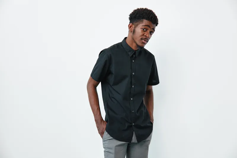 Young black man with dark skin wearing black shirt and gray pants standing with hands in pockets on white background. Casual style and confident pose in studio.
