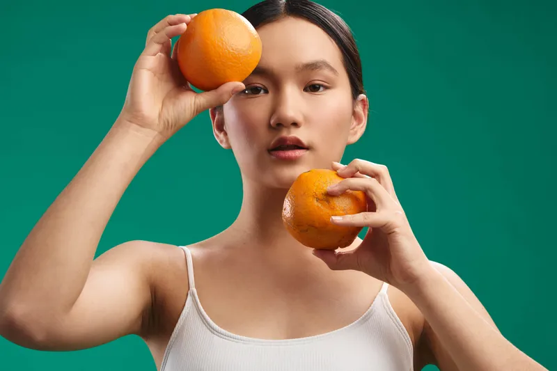 Asian woman holds oranges against a green backdrop. She wears a white tank top, gazes at the camera with a calm expression, creating a bright, vibrant still life mood.