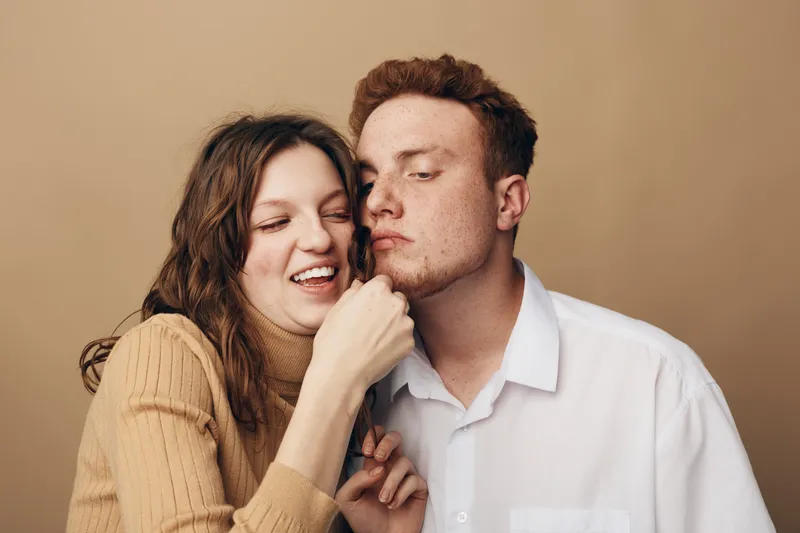 Happy young couple in casual clothes share a playful moment, woman smiling and touching man's face. Warm studio lighting creates a cozy and intimate atmosphere of affection and joy.