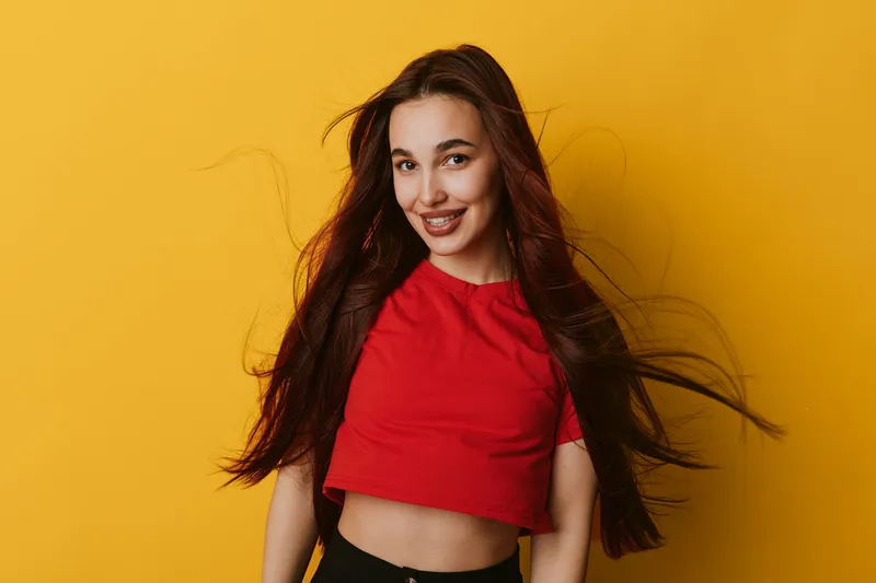 Young woman with long hair smiling and wearing red crop top on yellow background. Portrait of cheerful female with flowing hair and casual style for positive mood and fashion.
