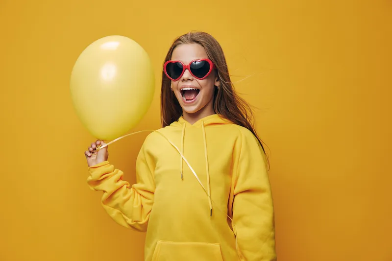 Girl with long hair wearing red heart-shaped sunglasses and yellow hoodie holding yellow balloon smiling with open mouth on yellow background. Happy, cheerful, joyful child portrait.