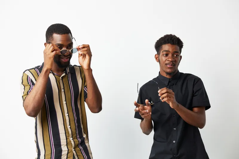 Two black men smiling and wearing sunglasses, showcasing stylish summer outfits and a confident demeanor against a clean white background.