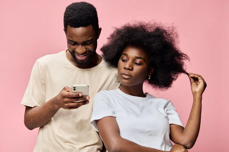 Young couple enjoying each other s company while looking at a smartphone against a pink background, showcasing casual fashion and playful emotions