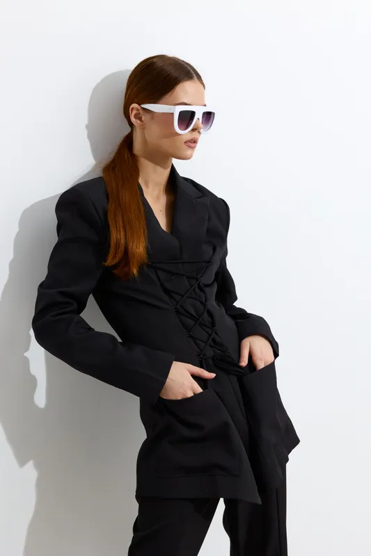 Fashion woman in stylish black outfit and white sunglasses posing confidently against white wall, showcasing modern beauty and sleek contemporary style.