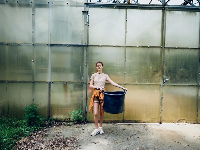 Woman in front of greenhouse holding a large black bucket, casual clothing and sneakers, gardening scene with plants and soil, outdoor agriculture and horticulture activity.