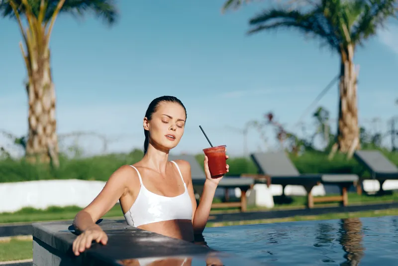 Relaxed woman in a white swimsuit lounges at a pool edge, holding a drink, palm trees and sunshine overhead, creating a summer relaxation moment.