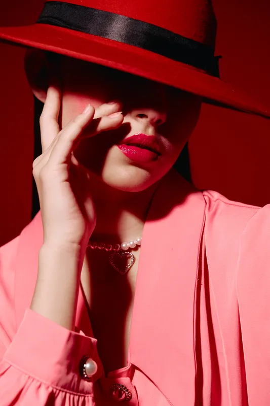 Woman in pink fashion wears wide-brimmed hat and bright lipstick, posing with hand near face. Bold coloring and shadows create a dramatic beauty concept for creative fashion.