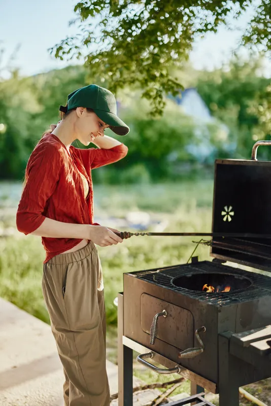 Young woman grilling outdoors in casual clothing, showcasing joy and relaxation amidst greenery, capturing the essence of summer gatherings and culinary joy