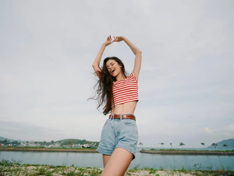 A joyful young woman stands outdoors with arms raised, wearing a red striped crop top and denim shorts, bright sky behind, capturing a moment of carefree summer energy.