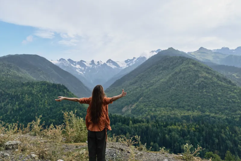 Person standing on a mountain overlook with arms wide, embracing vast valley and snowy peaks, conveying freedom, adventure, and connection with nature.