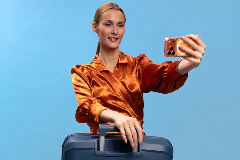 Young woman wearing shiny orange blouse takes selfie with smartphone while holding blue suitcase on blue background. Concept of travel, vacation, modern technology, and lifestyle.