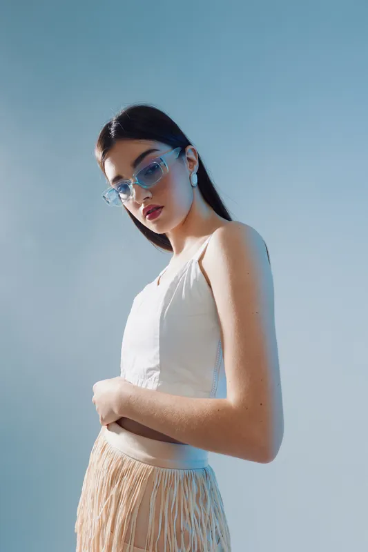 Young woman in white top and fringe skirt poses confidently wearing blue sunglasses against a clear sky. Concept of woman beauty fashion with modern style and attitude.