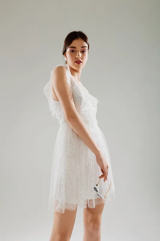 Young woman in a white lace dress poses confidently against a neutral background. Her makeup is natural, highlighting beauty and fashion concepts in a minimalist studio setting.