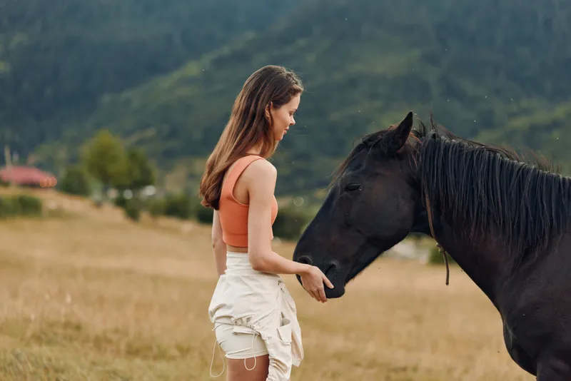 A young woman in an orange sleeveless top and light shorts stands in a rural field, gently touching a dark horse, creating a calm, pastoral moment.