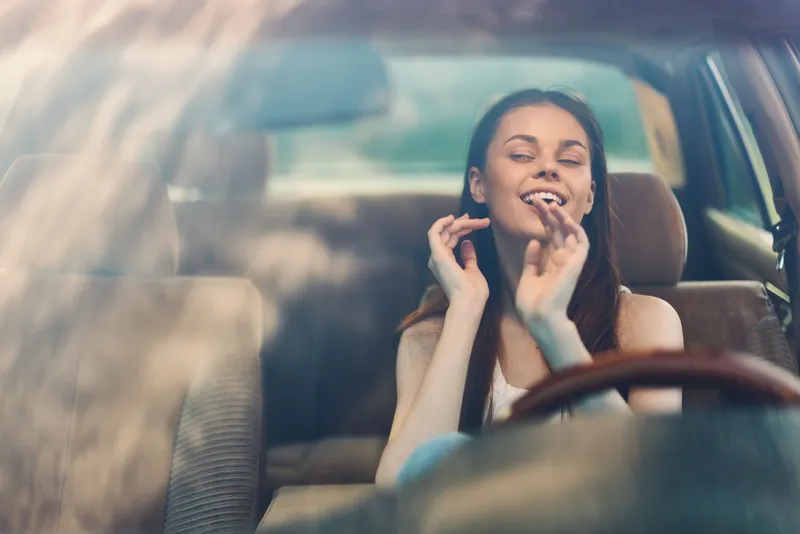Young woman enjoying music in a car, smiling brightly with carefree emotions, capturing the essence of joy and freedom on a sunny day.