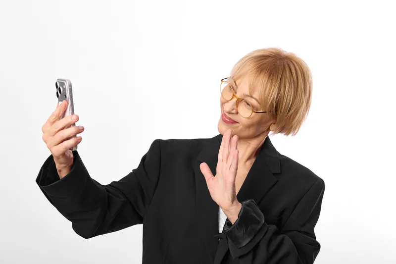 Confident mature woman with glasses in studio taking selfie with smartphone, making playful peace sign with hand, white background, close portrait, copy space