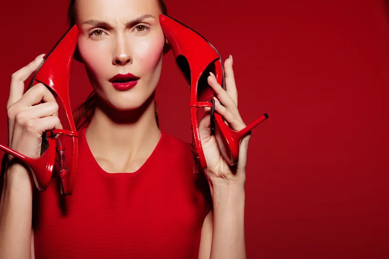 Woman in red dress holds red high heels near face against red background. Fashion concept with bold makeup and intense expression in studio lighting.