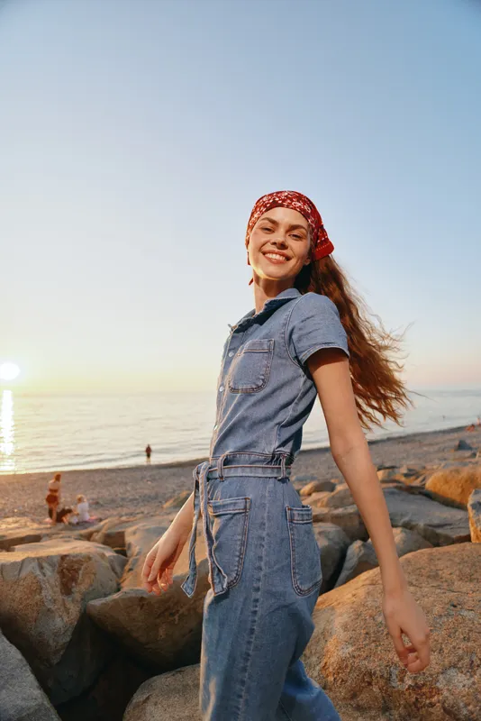Lifestyle woman in modern boho-western denim street style smiling by the beach at sunset with film color tones creating warm and natural mood.