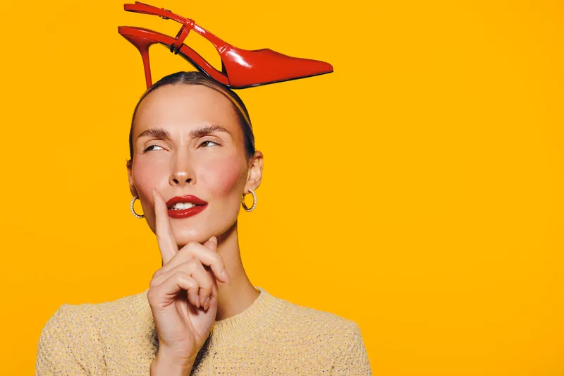 Stylish woman with red lipstick and earrings balances red high heel shoe on head against bright orange background. Concept for fashion, creativity, and playful expression.
