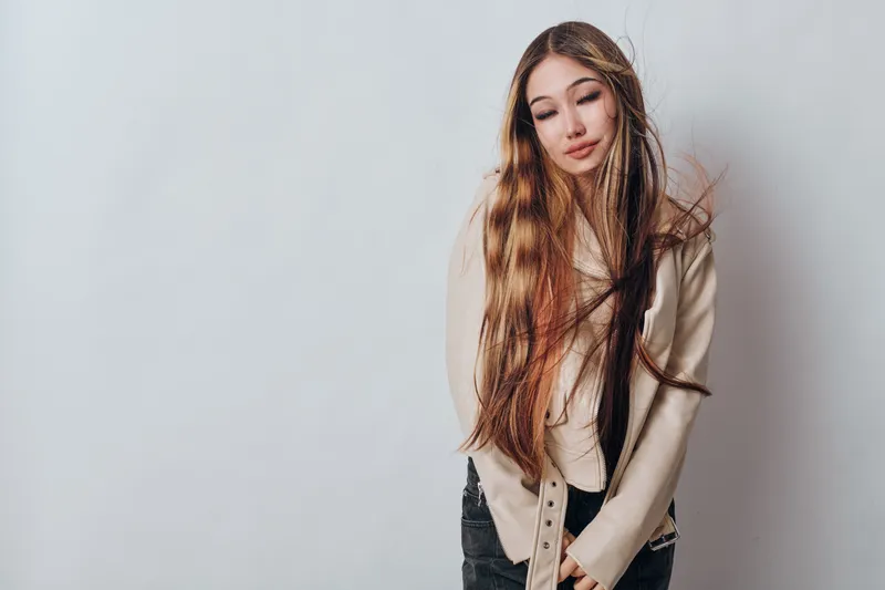 Young woman with long hair and neutral expression posing against light gray background, wearing casual beige sweater and black pants in studio photography.