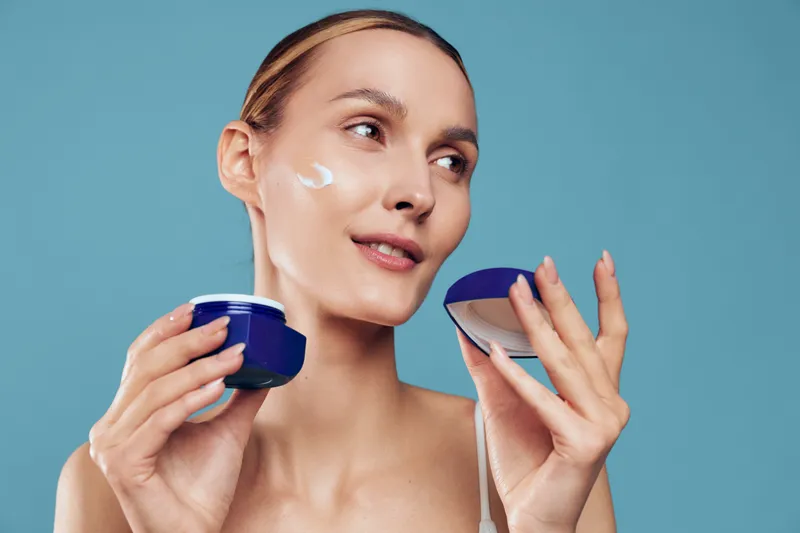 A young woman applies cream on her face while holding a jar of moisturizer against a blue background. The skincare concept focuses on beauty and self-care routines.