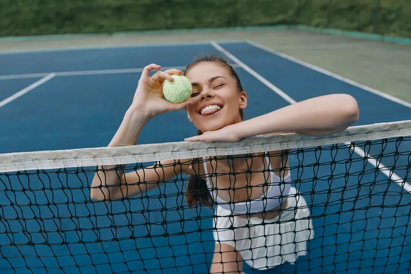 Joyful young female tennis player celebrating with a tennis ball on a sunny court, wearing sporty attire and showing playful emotions