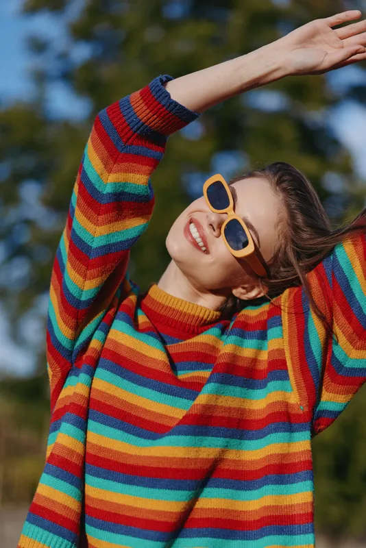 Young woman in her mid 20s wearing a colorful striped sweater and yellow sunglasses, smiling and joyful with arms raised outdoors in a sunlit park, casual outfit lifestyle fashion.