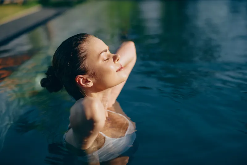 Relaxed woman in white swimsuit enjoying calm water, eyes closed, head tilted back, peaceful moment, natural light reflecting on skin, summer outdoor pool scene