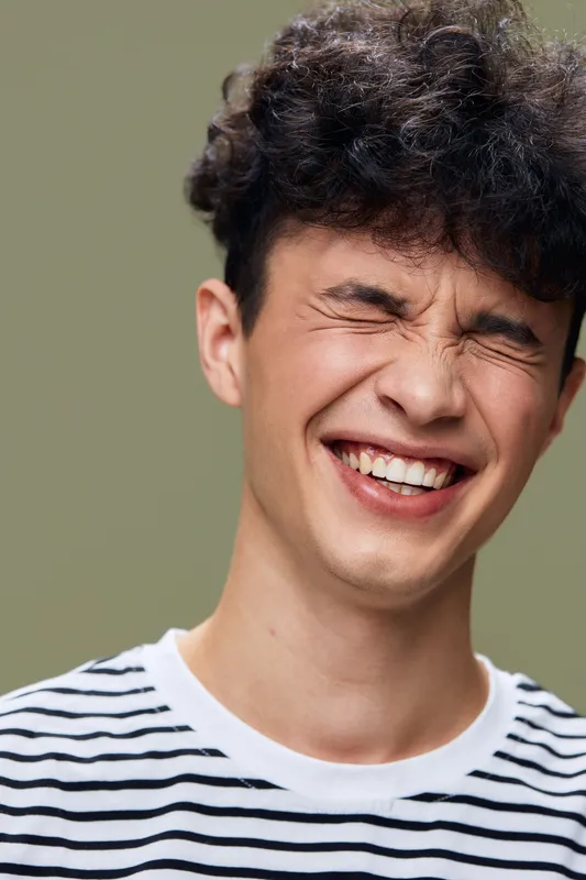 smiling man with curly hair in striped shirt laughing with closed eyes, natural joyful expression, studio portrait with muted green background