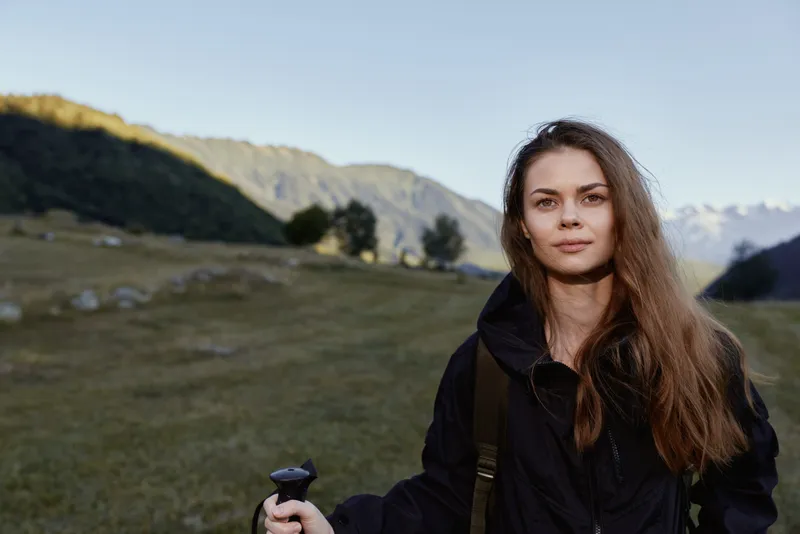 A cheerful female hiker stands in a scenic mountain meadow with a trekking pole in hand. She looks at the camera with calm confidence, enjoying nature and fresh air.