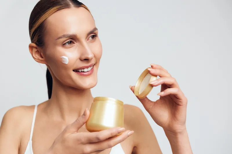 A smiling woman with clean skin holds an open jar of cream while applying moisturizer to her cheek in a bright studio. Concept for skincare, beauty, and self-care routine.