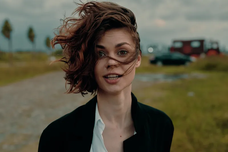 Woman outdoors with windy hair, light makeup and a black blazer, bright smile and relaxed pose along a gravel path in a breezy field during golden hour by the coast