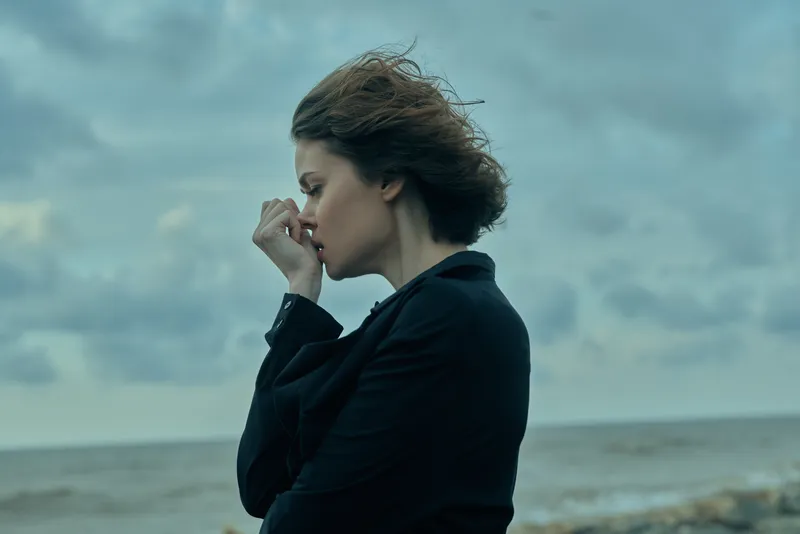 A contemplative woman in a dark blazer stands by a windy shoreline, hair swept by the breeze, hand near lips, evoking reflection and mood by the sea