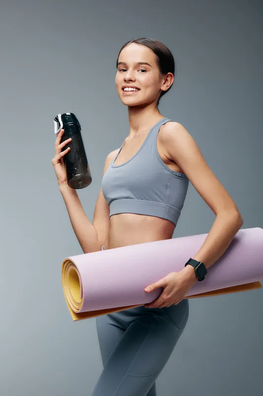 Female fitness enthusiast in a light blue athletic set holds a water bottle and a rolled yoga mat, ready for a workout in a clean studio with focused energy and training.