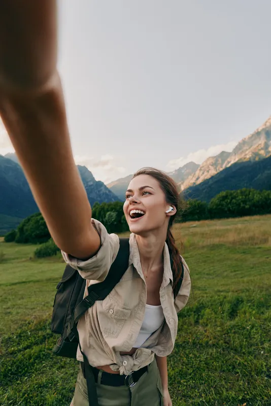 Young woman with braided hair wearing a beige shirt and backpack takes a selfie outdoors in a mountain meadow while smiling joyfully during a hiking trip.