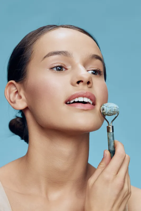 Young woman using jade roller for skincare, promoting healthy beauty routines and skincare rituals Blue background enhances freshness and tranquility