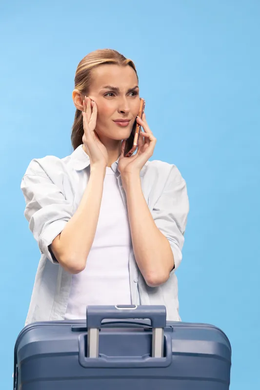 Young woman with suitcase talks on phone and looks worried or confused, standing against blue background. Concept of travel problems, communication, and uncertainty during trip.
