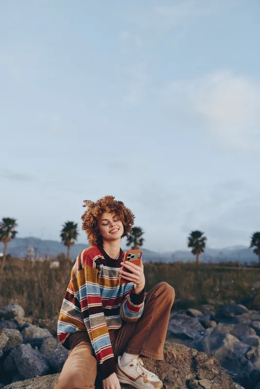 Woman outdoors lifestyle smartphone selfie casual sitting nature curly hair smiling, holding smartphone on rocks in desert landscape, palm trees distant, soft clouds overhead, relaxed travel