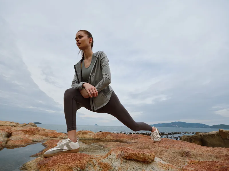 Young woman practicing yoga on rocky beach at sunrise, wearing athletic clothing, showcasing strength and balance while surrounded by nature