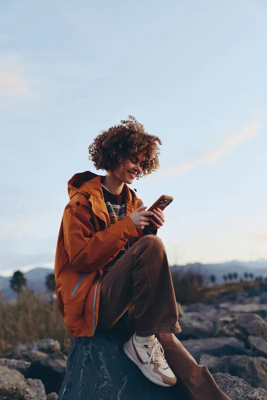 Outdoors lifestyle woman with curly hair, orange jacket, sits on rocks holding smartphone, smiling as she relaxes, mountain landscape behind, travel communication in evening light